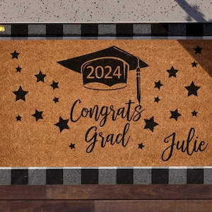 May include: A brown coir doormat with a black and white checkered border. The doormat has a black graduation cap with the year 2024 on it, the words "Congrats Grad" and the name "Julie" written in black.