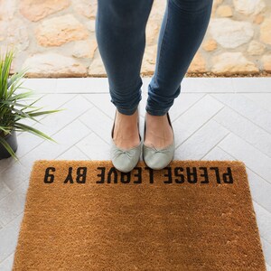May include: A brown doormat with the text "PLEASE LEAVE BY 6" in black lettering. A person is standing on the doormat wearing light blue jeans and grey ballet flats.