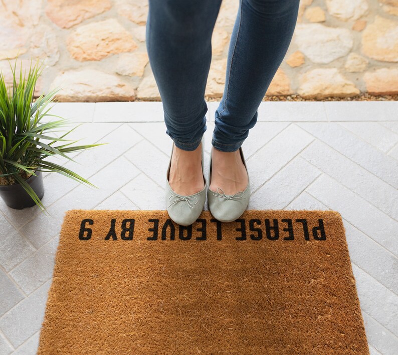May include: A brown doormat with the text "PLEASE LEAVE BY 6" in black lettering. A person is standing on the doormat wearing light blue jeans and grey ballet flats.