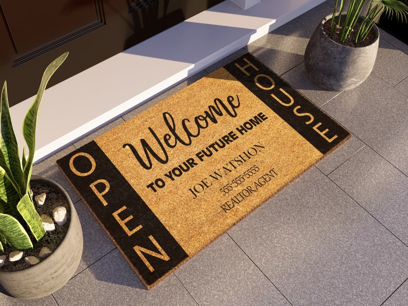 Op de afbeelding: Een bruine kokosmat met zwarte letters die "Open House Welcome to your future home Joe Wattshon 555-555-5555 Realtor Agent" zeggen.