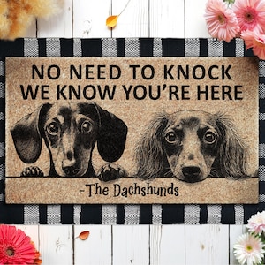 May include: A rectangular doormat with the text "NO NEED TO KNOCK WE KNOW YOU'RE HERE" and "-The Dachshunds". The mat features illustrations of two dachshunds. The mat is brown with a black and white checkered border.