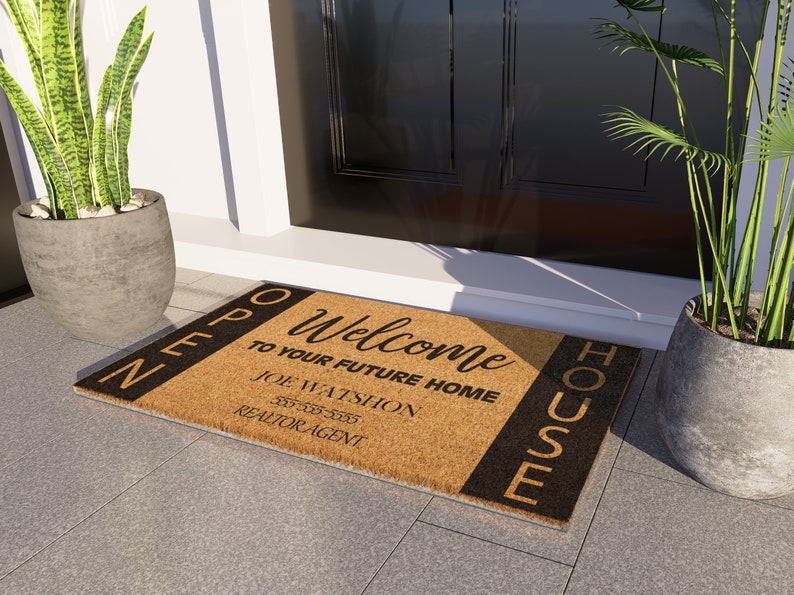 Op de afbeelding: Een bruine kokosvezel deurmat met zwarte strepen en de tekst "Welkom in uw toekomstige huis" en "Open huis" met de naam en het telefoonnummer van een makelaar.