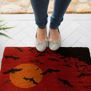 May include: A red and black doormat with a large orange moon and black bats flying around it.