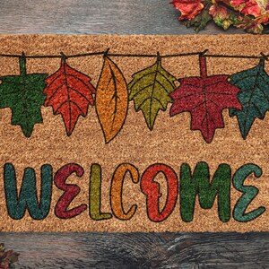 May include: A brown coir doormat with the word "WELCOME" in colorful letters. The letters are outlined in black and have a fall-themed design. A string of colorful leaves hangs above the word.