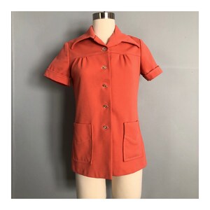 May include: A short-sleeved, coral-colored button-down shirt with a collar and two patch pockets. The shirt is on a mannequin.