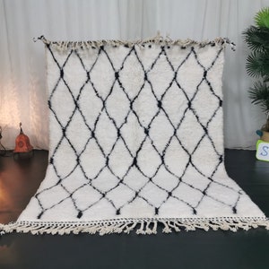 May include: White and black Moroccan rug with a diamond pattern and fringe. The rug is laid out on a dark floor.