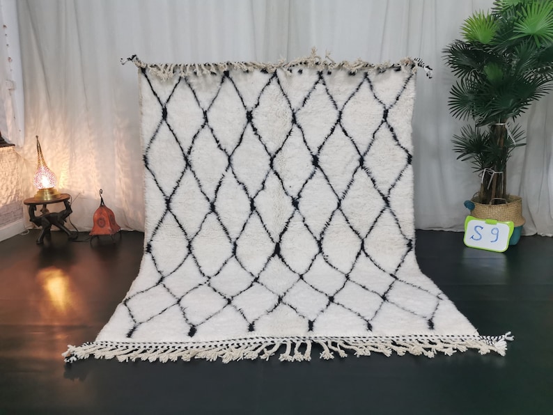 May include: White and black Moroccan rug with a diamond pattern and fringe. The rug is laid out on a dark floor.