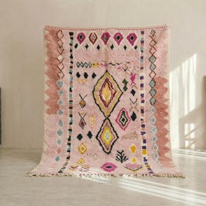 Puede incluir: Una gran alfombra marroquí rosa con patrones geométricos en negro, amarillo y azul. La alfombra presenta formas de diamantes y rectangulares, con un borde con flecos. Una silla decorativa y una fuente de mosaico están en el fondo.
