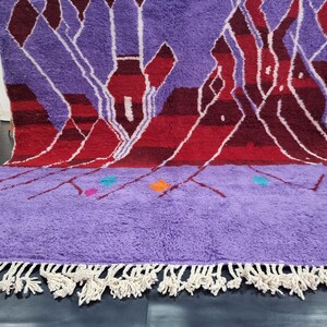 May include: A hand-woven Moroccan rug with a purple background and a geometric pattern in shades of red, white, and orange. The rug has a fringe border.