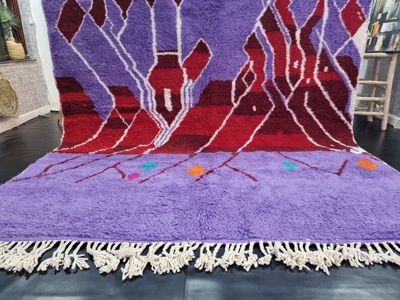 May include: A hand-woven Moroccan rug with a purple background and a geometric pattern in shades of red, white, and orange. The rug has a fringe border.