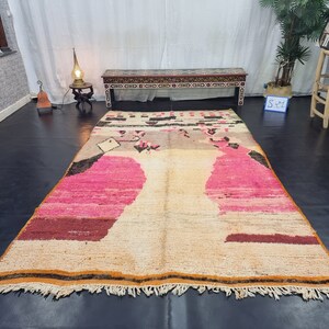 May include: A large, rectangular, hand-woven rug with a cream background and pink, brown, and black geometric patterns. The rug has a fringe border.