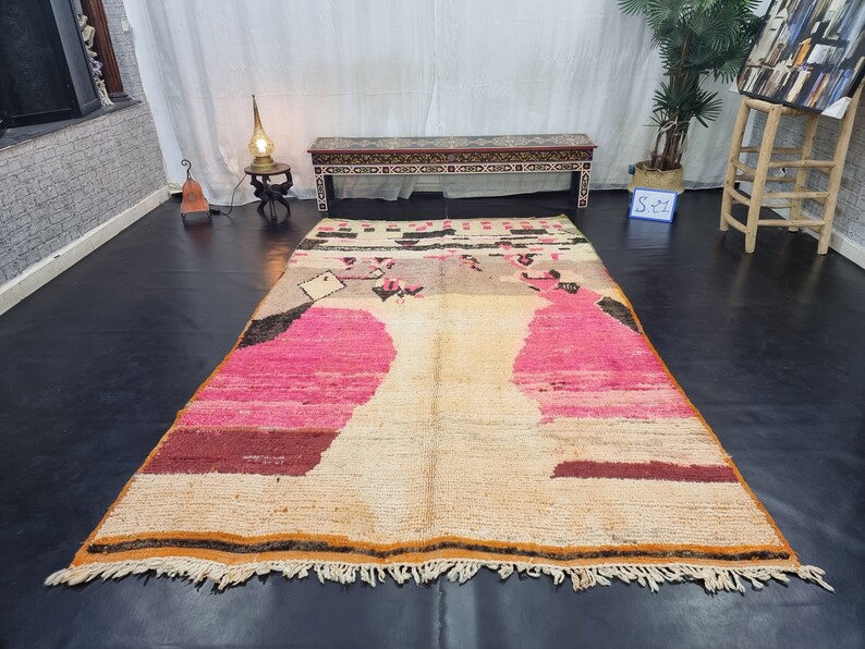 May include: A large, rectangular, hand-woven rug with a cream background and pink, brown, and black geometric patterns. The rug has a fringe border.