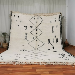 May include: A white Moroccan rug with black geometric patterns and fringe. The rug has a traditional design with a central diamond shape and smaller diamonds surrounding it.