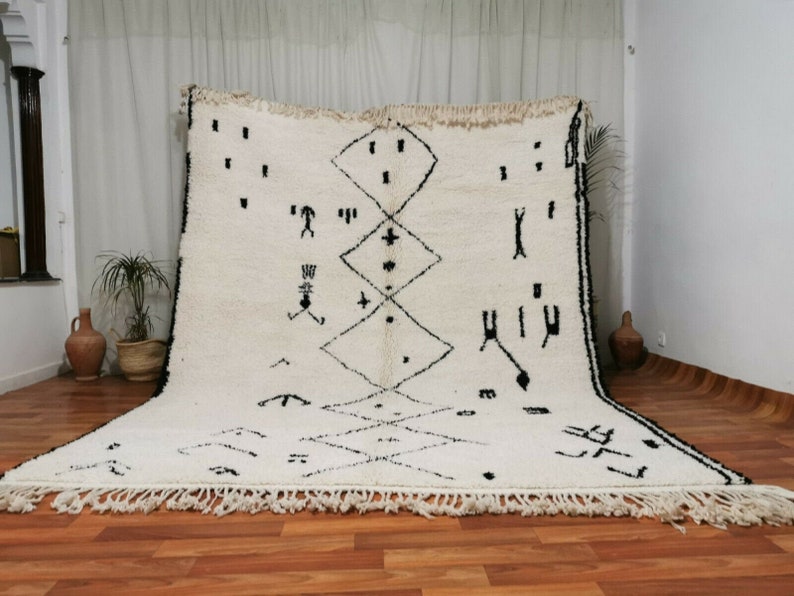 May include: A white Moroccan rug with black geometric patterns and fringe. The rug has a traditional design with a central diamond shape and smaller diamonds surrounding it.