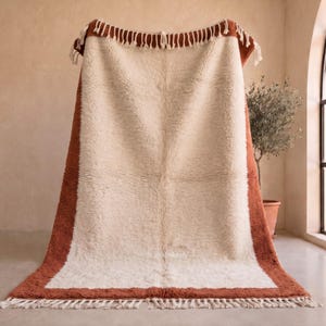 Puede incluir: Una alfombra rectangular que presenta un gran panel central de color crema enmarcado por un borde ancho y sólido de color terracota. La alfombra tiene una textura de pelo largo y grueso, y está rematada con largos flecos color crema en los extremos.