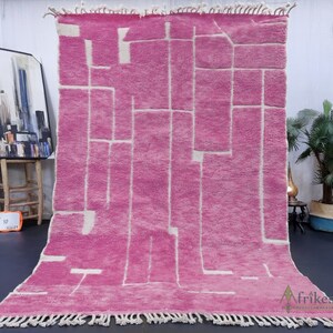 May include: A pink and white geometric patterned rug with fringe on the edges. The rug is laid out on a dark floor with a painting and a plant in the background.
