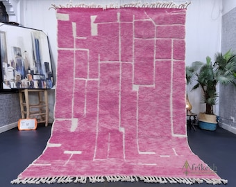 May include: A pink and white geometric patterned rug with fringe on the edges. The rug is laid out on a dark floor with a painting and a plant in the background.