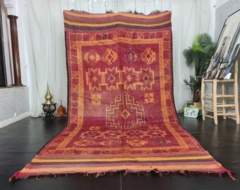 May include: A large, red and orange Moroccan rug with intricate geometric patterns. The rug has a fringe border and is woven with wool.