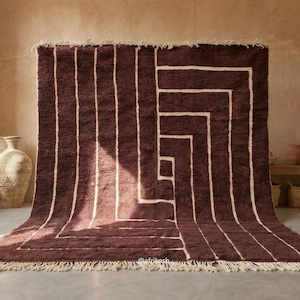 May include: A large, rectangular, deep burgundy area rug featuring a geometric pattern of thick, cream-colored lines that form a spiral design. The rug has a thick, textured pile and is finished with a cream-colored fringe along the bottom edge.