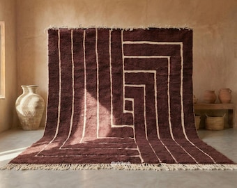 May include: A large, rectangular, deep burgundy area rug featuring a geometric pattern of thick, cream-colored lines that form a spiral design. The rug has a thick, textured pile and is finished with a cream-colored fringe along the bottom edge.