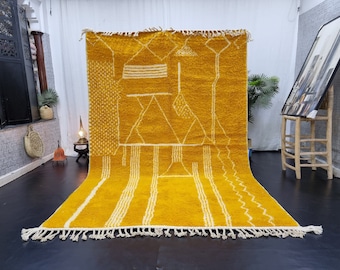 May include: A large, rectangular, mustard yellow rug featuring a white geometric pattern of lines, triangles, and dots. The rug has a thick, textured pile and is finished with white knotted fringe along the top and bottom edges.