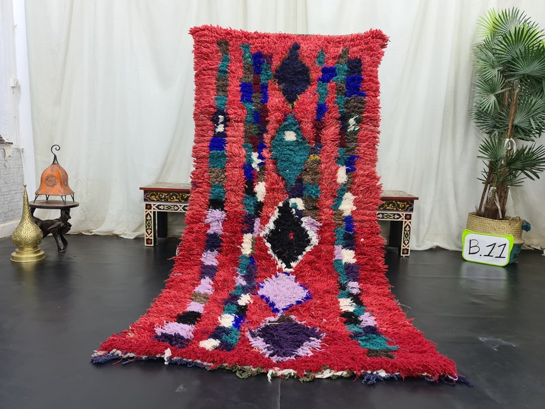 May include: A red, blue, green, and white Moroccan rug with a geometric pattern. The rug is made of wool and has a shaggy texture. The rug is approximately 7 feet long and 3 feet wide.