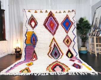 May include: A white Moroccan rug with a colourful diamond pattern. The rug has a fringe border and is made of wool.