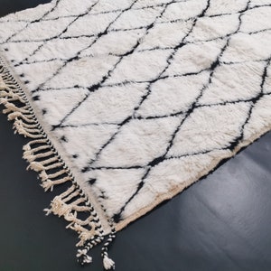 May include: A white and black Moroccan rug with a diamond pattern and fringe. The rug is made of wool and has a soft, fluffy texture.