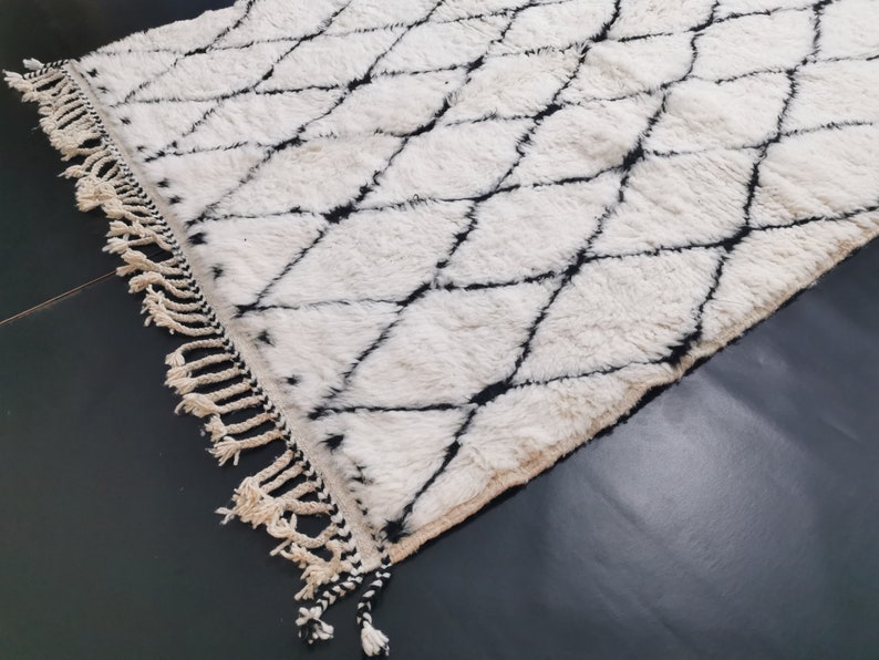 May include: A white and black Moroccan rug with a diamond pattern and fringe. The rug is made of wool and has a soft, fluffy texture.