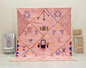 May include: A large, pink Moroccan rug with geometric patterns and tribal designs in various colors. The rug features diamond shapes, triangles, and other symbols, with a fringe along the bottom edge. The rug is displayed against a neutral background.