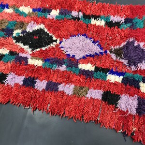 May include: A colorful, hand-woven rug with a red background and a variety of geometric patterns in shades of purple, blue, green, and white.