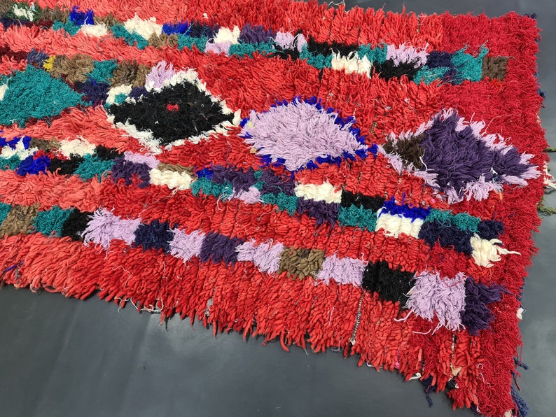 May include: A colorful, hand-woven rug with a red background and a variety of geometric patterns in shades of purple, blue, green, and white.