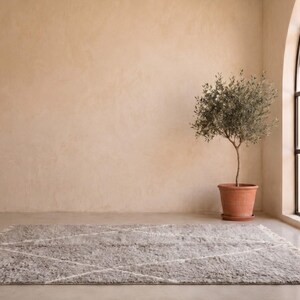 May include: A rectangular area rug featuring a light grey, shaggy texture with a subtle white geometric diamond pattern and fringed edges. The rug is positioned on a smooth floor, providing a soft, textured surface with a neutral colour palette.