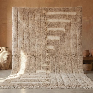 May include: A large, beige rug with a geometric design. The rug has a shaggy texture and a rectangular shape. The design features vertical stripes and a series of horizontal and diagonal lines in a lighter shade of beige. The rug is displayed in a room with neutral tones.