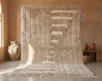 Puede incluir: Una alfombra beige grande con un diseño geométrico. La alfombra tiene una textura peluda y una forma rectangular. El diseño presenta rayas verticales y una serie de líneas horizontales y diagonales en un tono beige más claro. La alfombra se exhibe en una habitación con tonos neutros.