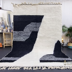 May include: A white, black, and gray Moroccan rug with a geometric design. The rug has a fringe border and is made of wool.