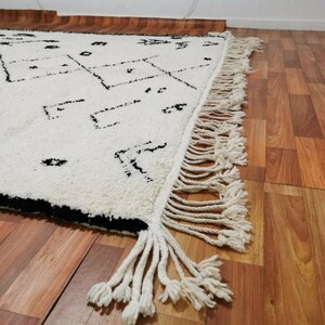 May include: A white and black Moroccan rug with a geometric pattern and fringe. The rug is laid out on a wooden floor.