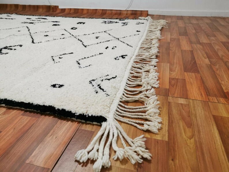 May include: A white and black Moroccan rug with a geometric pattern and fringe. The rug is laid out on a wooden floor.