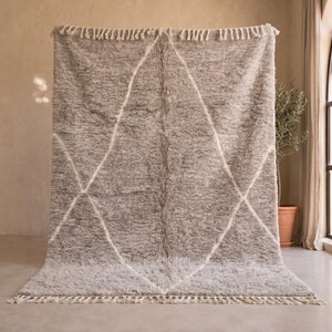 May include: A rectangular, high-pile area rug in a soft grey tone, featuring a white diamond-shaped geometric pattern across its surface. The rug is finished with short, cream-coloured fringe tassels along the top and bottom edges.