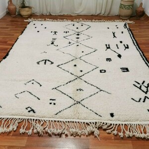 May include: A white Moroccan rug with black geometric patterns and fringe. The rug has a traditional design with diamond shapes and abstract symbols.
