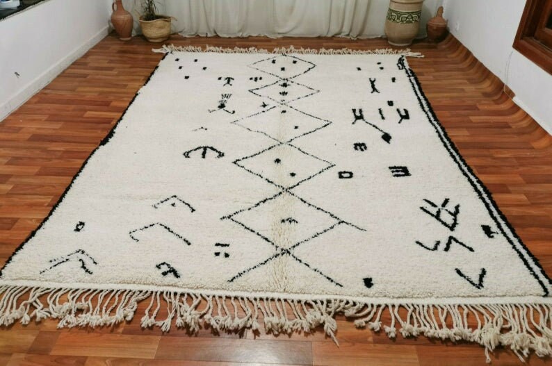 May include: A white Moroccan rug with black geometric patterns and fringe. The rug has a traditional design with diamond shapes and abstract symbols.