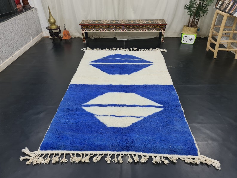 Puede incluir: Una alfombra blanca y azul con un borde de flecos. La alfombra tiene dos formas abstractas blancas que se asemejan a labios sobre un fondo azul.