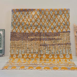 Può includere: Un tappeto tessuto a mano con un motivo geometrico in tonalità di giallo, grigio e viola. Il tappeto ha un bordo a frangia ed è fatto di lana.