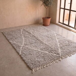 May include: A rectangular area rug with a thick, shaggy pile in a light grey tone. The rug features a white geometric diamond pattern outlined in thin lines and is finished with decorative knotted fringe along the two shorter edges.