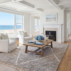May include: A living room featuring two white upholstered armchairs with patterned cushions, a wooden coffee table with blue flowers and books, a grey shag rug with a white diamond pattern, and a fireplace with a white mantel and ocean view.