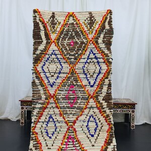 May include: A white, brown, red, blue, and pink Moroccan rug with a diamond pattern. The rug is handwoven and has a fringed edge.