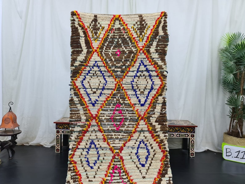May include: A white, brown, red, blue, and pink Moroccan rug with a diamond pattern. The rug is handwoven and has a fringed edge.