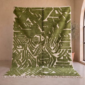 May include: A rectangular area rug featuring a high-pile texture with an olive green and cream geometric pattern. The design consists of abstract, angular shapes and lines. The rug includes a fringed edge at the bottom and the text @afrikesh.