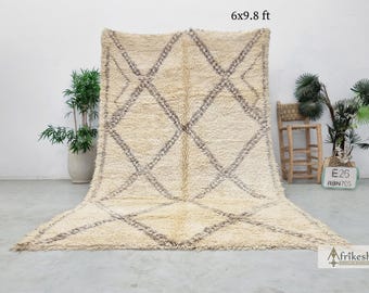 May include: A large, off-white, hand-woven Moroccan rug with a diamond pattern in a darker shade of beige. The rug is 6x9.8 feet and is shown on a light gray floor.