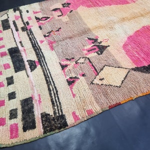May include: A vintage Moroccan rug with a geometric pattern in shades of pink, black, and white. The rug has a distressed look and is made of wool.